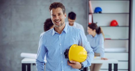 smiling-male-senior-architect-holding-helmet-hands-while-standing-office